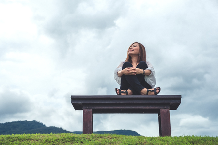 A happy asian woman sitting on a wooden bench in the park with blue sky backgroundの写真素材