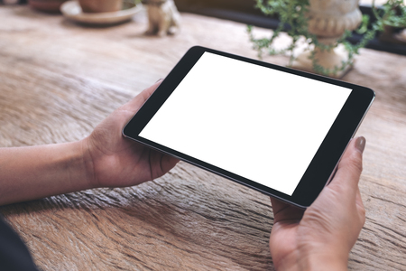 Mockup image of hands holding black tablet pc with blank white screen on vintage wooden tableの写真素材