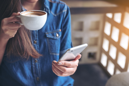 Closeup image of a woman holding , using and touching a smart phone while drinking coffeeの写真素材