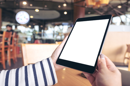 Mockup image of hands holding black tablet pc with blank white desktop screen on wooden table in cafeの写真素材