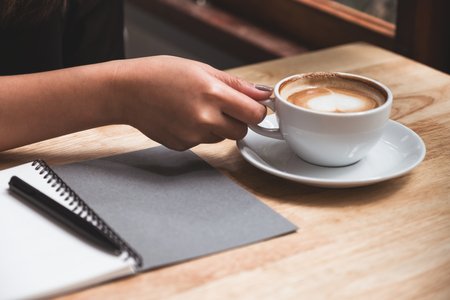 Closeup image of a woman's hand writing down on a white blank notebook while drinking coffee on wooden tableの写真素材