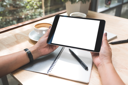 Mockup image of a woman's hands holding black tablet pc with white blank screen with notebook and coffee cup on tableの写真素材