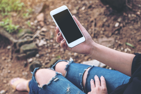 Mockup image of woman holding white mobile phone with blank desktop screen while sitting on the ground outdoorの写真素材