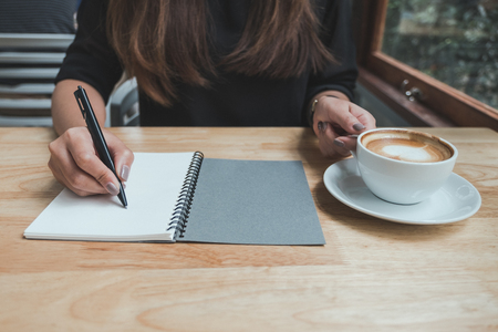 Closeup image of a woman's hand writing down on a white blank notebook while drinking coffee on wooden tableの写真素材