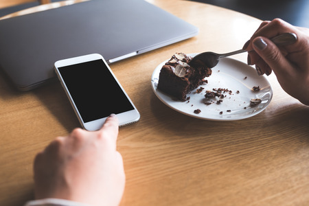 Mockup image of a woman pointing at white mobile phone with blank black desktop screen while enjoy eating brownie cake in cafeの写真素材