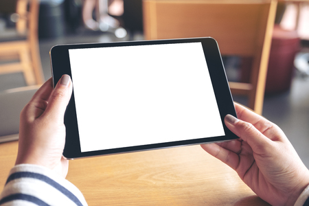 Mockup image of hands holding black tablet pc with blank white desktop screen on wooden table in cafeの写真素材