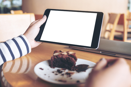 Mockup image of woman holding black tablet pc with blank white desktop screen while enjoy eating brownie cake in cafeの写真素材