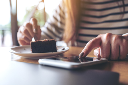 Closeup image of a woman using and touching a smart phone while eating brownie cake in cafeの写真素材