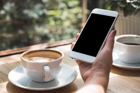 Mockup image of hand holding white mobile phone with blank black desktop screen with coffee cup on wooden table in cafeの写真素材