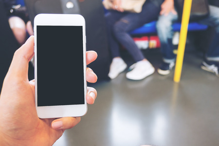 Mockup image of hand holding white mobile phone with blank black screen in subway with many people in backgroundの写真素材