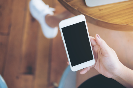 Mockup image of hands holding white mobile phone with blank black screen on thigh with silver colour laptop on table and wooden floor backgroundの写真素材