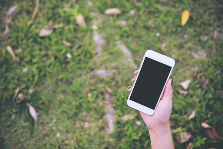 Mockup image of a hand holding white mobile phone with blank black screen in green garden floor backgroundの写真素材