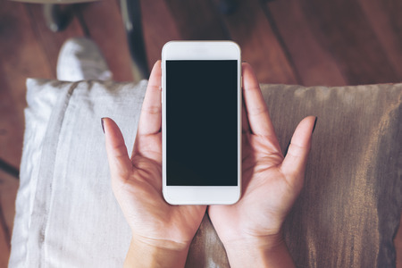 Mockup image of hands holding white mobile phone with blank black screen on pillow with wooden floor backgroundの写真素材