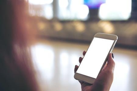 Mockup image of a woman holding and using white mobile phone with blank white screen while standing and waiting for baggage claim in the airportの写真素材