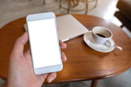 Mockup image of hand holding white mobile phone with blank desktop screen with coffee cup and laptop on tableの写真素材