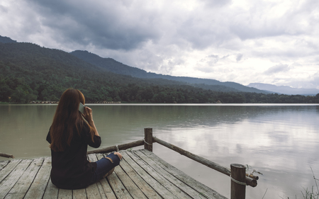 Closeup image of a woman sitting and talking on mobile phone on an old wooden pier by the river with sky and mountain backgroundの写真素材