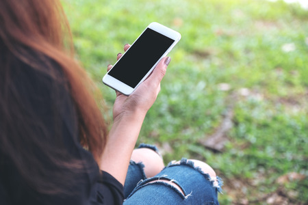 Mockup image of woman holding white mobile phone with blank black desktop screen while sitting on green yardの写真素材