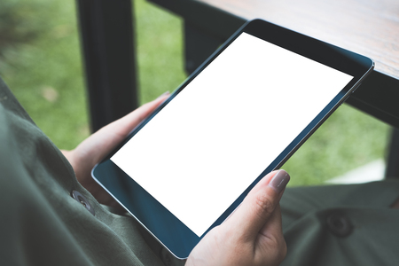 Mockup image of woman's hands holding black tablet pc with white blank screen while sitting in cafe with green nature backgroundの写真素材