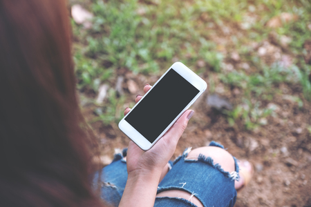 Mockup image of woman holding white mobile phone with blank black desktop screen while sitting on green yardの写真素材