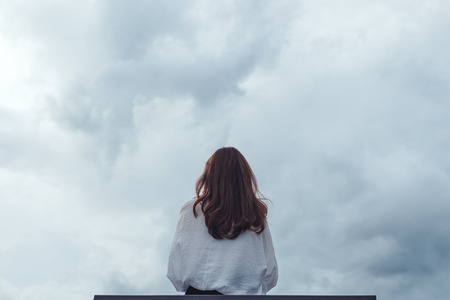 A woman sitting alone on a wooden bench in the park with cloudy and gloomy sky backgroundの写真素材