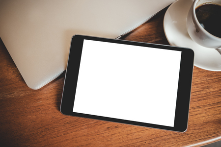 Top view mockup image of a black tablet pc with blank desktop white screen with laptop and coffee cup on wooden tableの写真素材