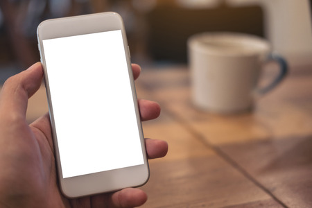 Mockup image of hand holding white mobile phone with blank desktop screen with coffee cup on wooden table in cafeの写真素材