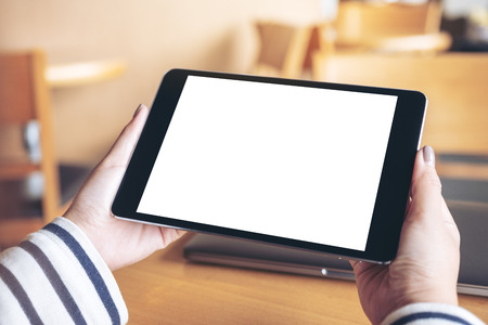Mockup image of hands holding black tablet pc with blank white desktop screen on wooden table in cafeの写真素材