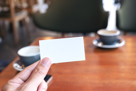 A hand holding white empty business card with two coffee cups on wooden table in cafeの写真素材