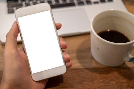 Mockup image of hand holding white mobile phone with blank desktop screen with coffee cup and laptop on wooden table in cafeの写真素材