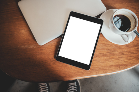 Top view mockup image of a black tablet pc with blank desktop white screen with laptop and coffee cup on wooden tableの写真素材