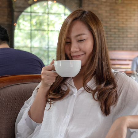 Closeup image of a beautiful Asian woman holding and drinking hot coffee with feeling good in cafeの写真素材