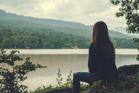 Closeup image of an asian woman sitting alone by the river with sky and mountain backgroundの写真素材