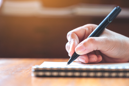 Closeup image of a hand writing down on a white blank notebook on wooden tableの写真素材