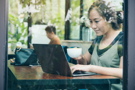 Closeup image of an asian business woman's working and using laptop on wooden table while drinking coffee in cafeの写真素材