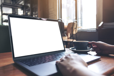 Mockup image of a woman using laptop with blank white desktop screen while drinking hot coffee on wooden table in cafeの写真素材