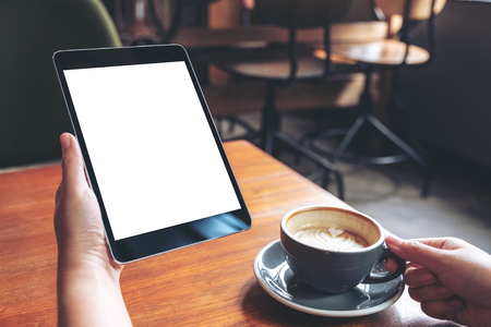 Mockup image of woman's hands holding black tablet pc with blank white screen with coffee cup on wooden table in cafeの写真素材