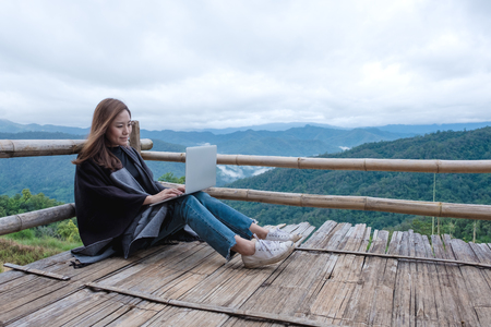 Closeup image of a woman working and typing on laptop while sitting on wooden balcony with green mountains on foggy day with blue sky backgroundの写真素材