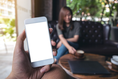 Mockup image of hand holding white mobile phone with blank screen in cafe and woman in backgroundの写真素材