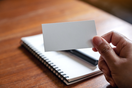A hand holding white empty business card with notebook on wooden tableの写真素材