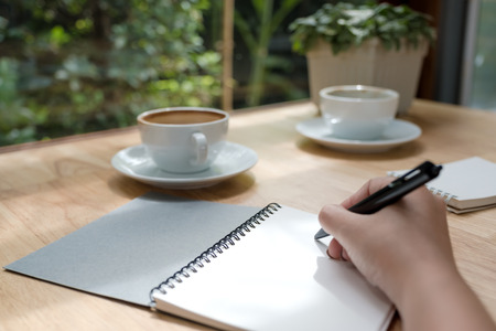 Closeup image of a woman writing down on a white blank notebook with coffee cup on wooden tableの写真素材