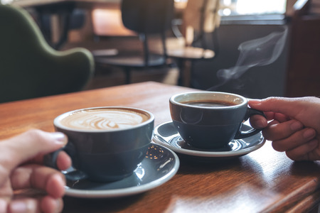 Closeup image of two people's hands holding coffee and hot chocolate cups on wooden table in cafeの写真素材