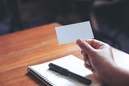 A hand holding white empty business card with notebook on wooden tableの写真素材