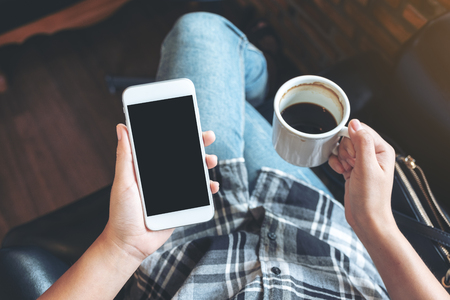 Top view mockup image of woman's hands holding white mobile phone with blank black desktop screen while drinking coffee in cafeの写真素材