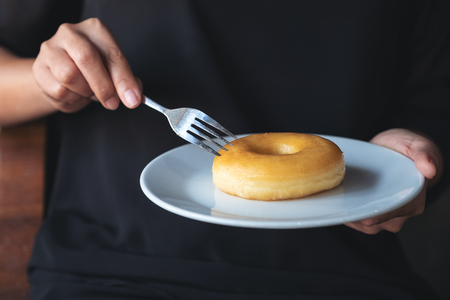 A woman's hands holding and cutting a piece of donut in a white plateの写真素材