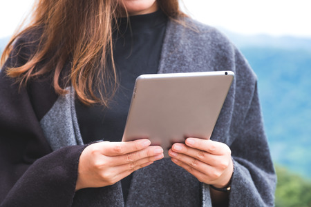Closeup image of a woman holding and using tablet pc with green mountains on foggy day with blue sky backgroundの写真素材