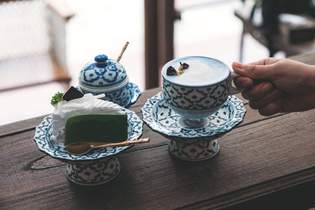 A hand holding a cup of hot milk with a piece of cake in an antique Thai traditional benjarong style containersの写真素材