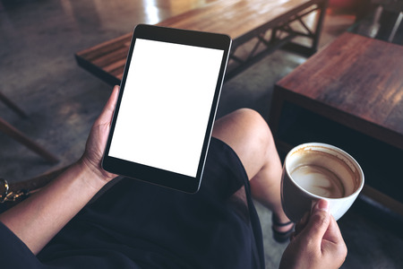 Mockup image of woman's hands holding black tablet pc with blank white desktop screen while sitting and drinking coffee in cafeの写真素材