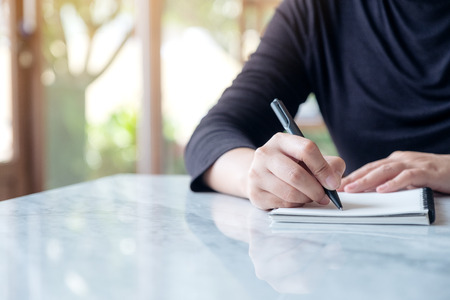 Closeup image of a woman writing on blank notebook on table with green nature backgroundの写真素材
