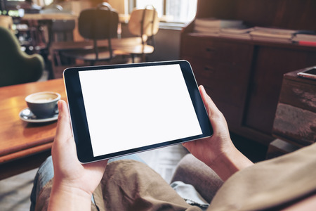 Mockup image of hands holding black tablet pc with blank white screen with coffee cup on wooden table in cafeの写真素材