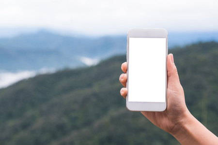 Mockup image of a hand holding and showing white smart phone with blank desktop screen in outdoor with blur green mountains backgroundの写真素材